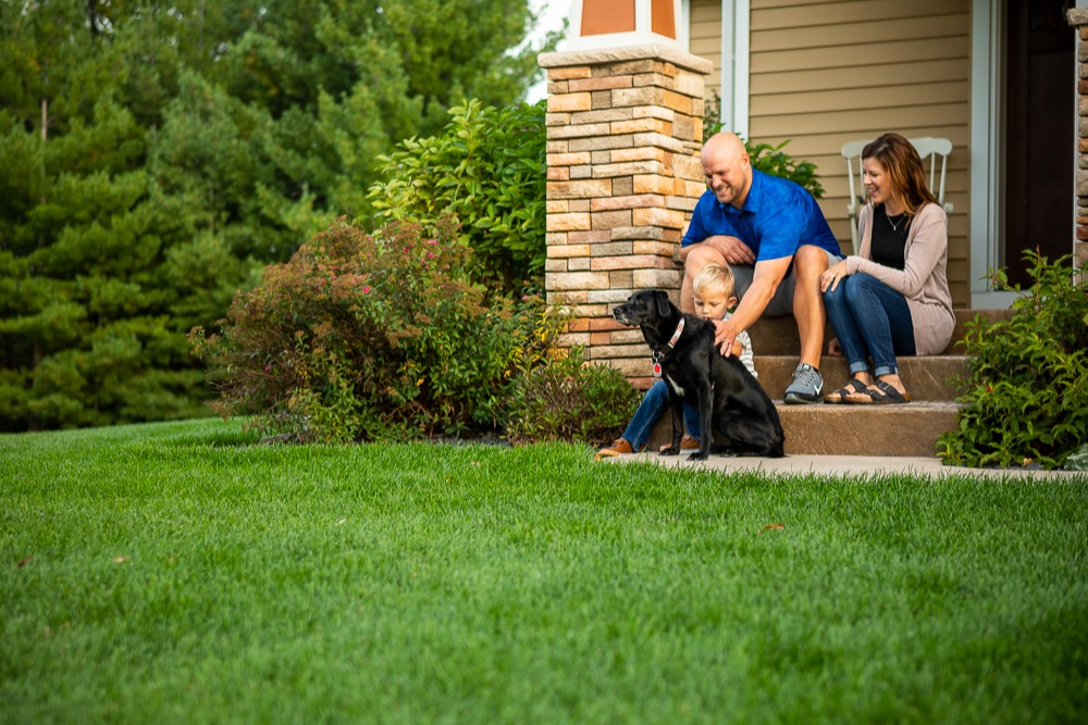 family sits on porch in front of green grass and plantings